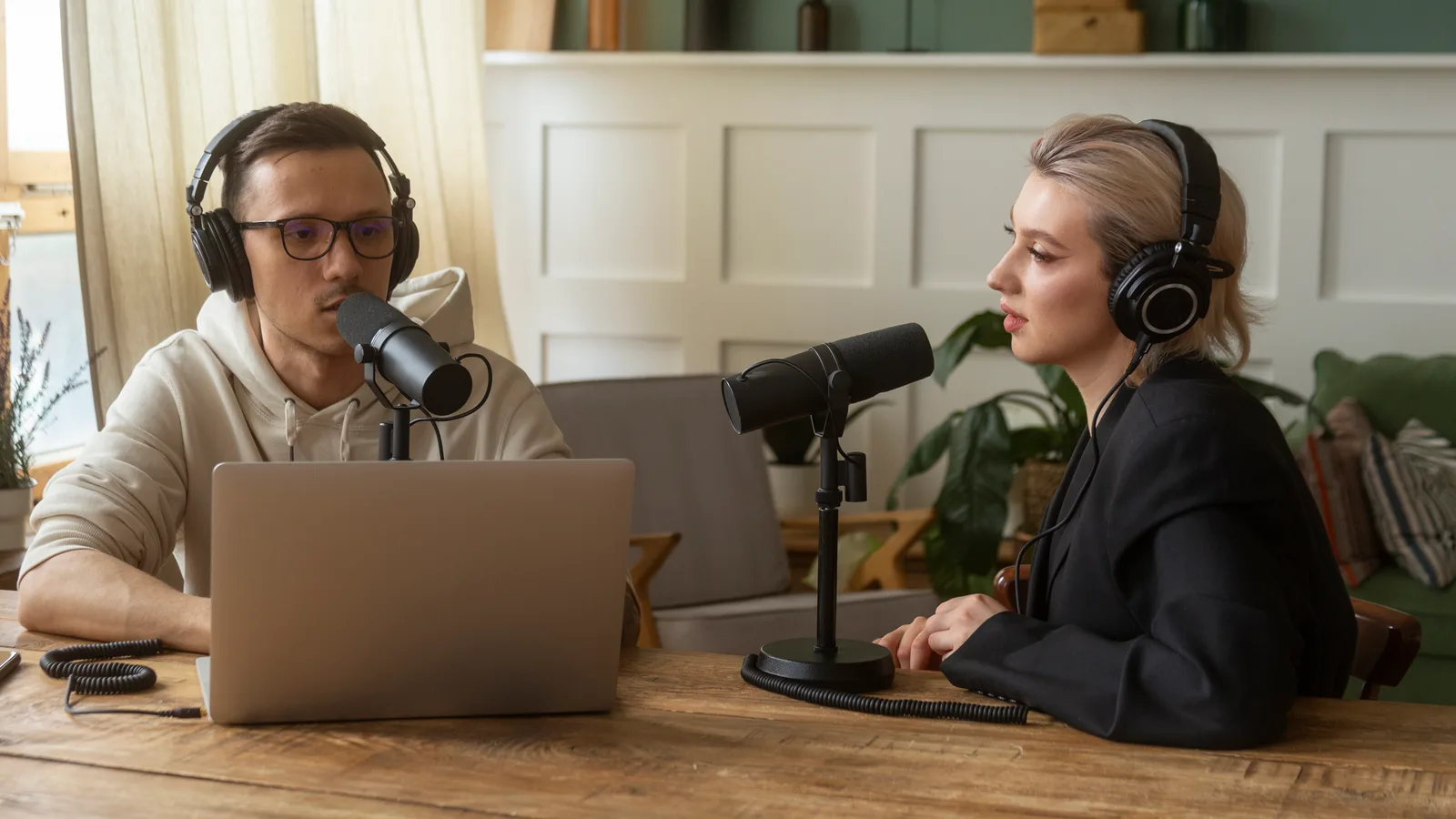 Wide podcast shot: two hosts seated at opposite ends of a wooden table, each behind a microphone