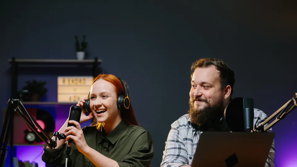 Wide podcast shot: two hosts at a studio desk, one laughing into a handheld microphone on the left, the other behind a laptop on the right
